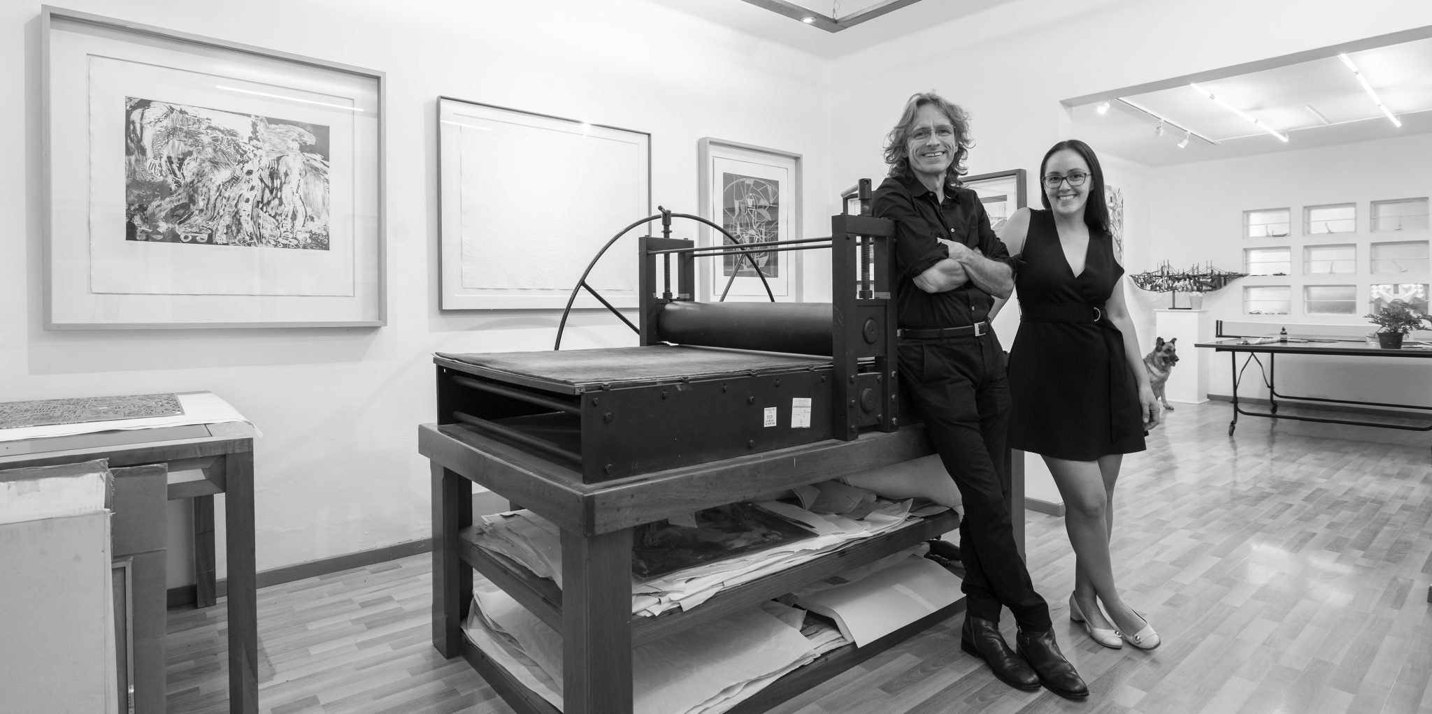 Two people standing next to a large printing press in an art gallery setting.