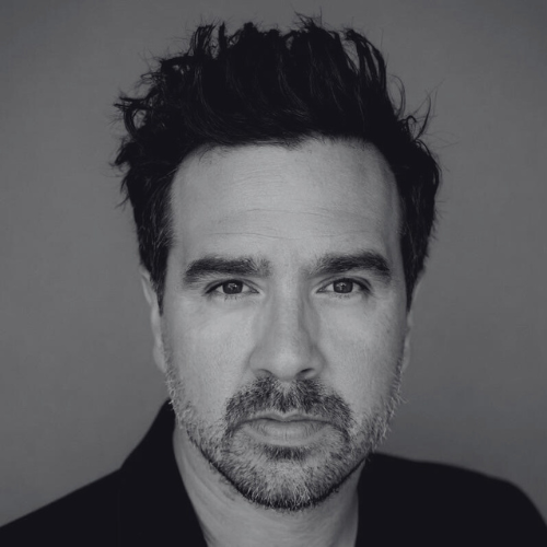 Black and white portrait of a man with spiky hair and beard against a plain background